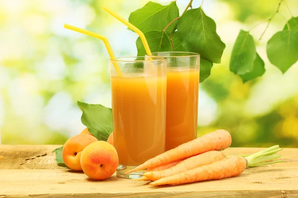 Glasses of carrot and apricot juice on wooden table on green background