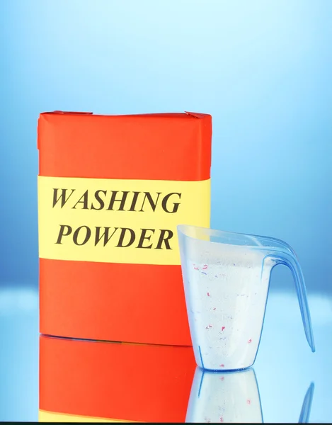 Box of washing powder with blue measuring cup, on blue background close-up
