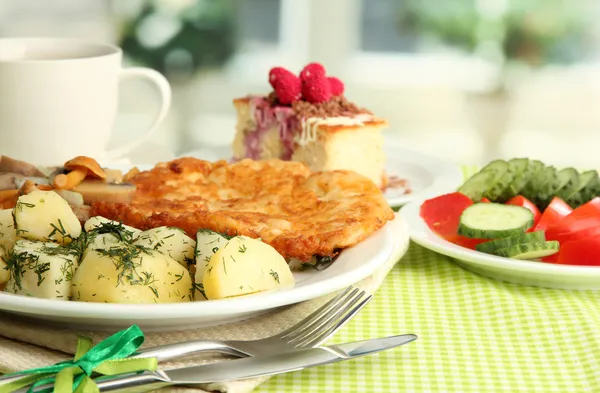 Roast chicken cutlet with boiled potatoes and cucumbers, cup of tea and dessert on green table cloth in cafe interior