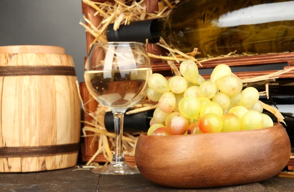 Wooden case with wine bottles, barrel, wineglass and grape on wooden table on grey background