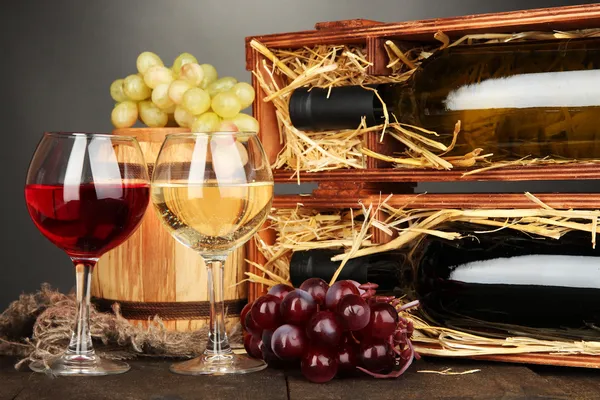 Wooden case with wine bottles, barrel, wineglasses and grape on wooden table on grey background