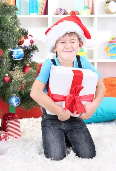 Little boy in Santa hat sits near Christmas tree with gift in hands