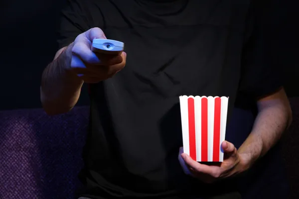 Man hand holding a TV remote control and popcorn, on dark background