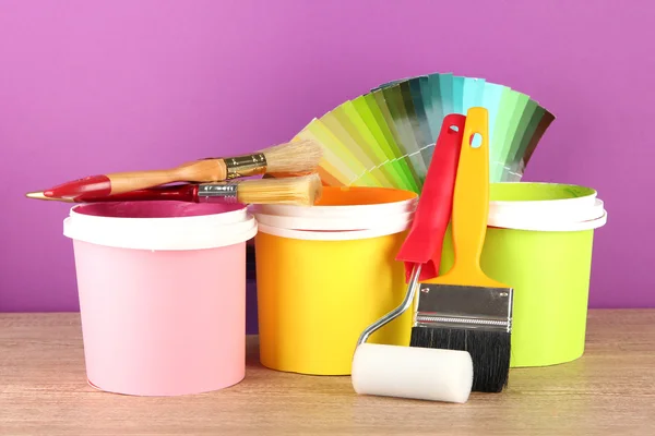 Paint pots, paintbrushes and coloured swatches on wooden table on purple background