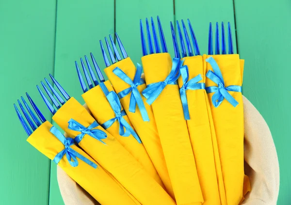 Blue plastic forks wrapped in yellow paper napkins, in basket, on color wooden background