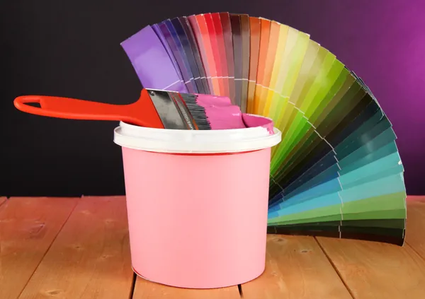Paint pot, paintbrush and coloured swatches on wooden table on dark purple background