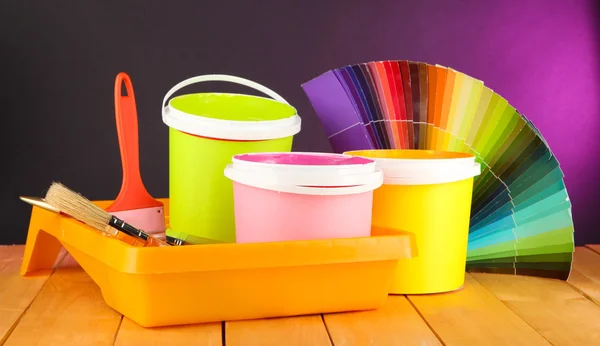 Paint pots, paintbrushes and coloured swatches on wooden table on dark purple background