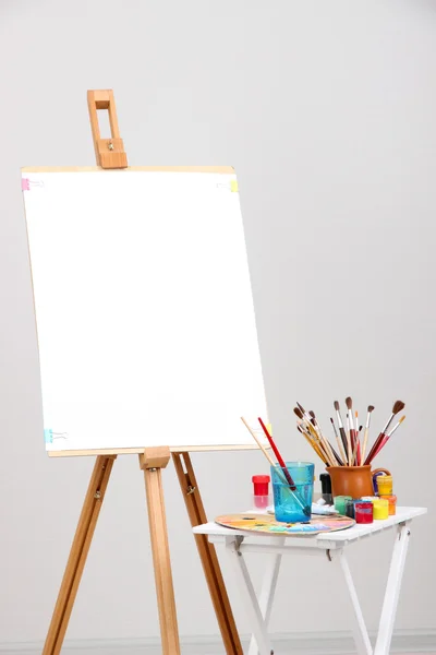 Wooden easel with clean paper and art supplies in room