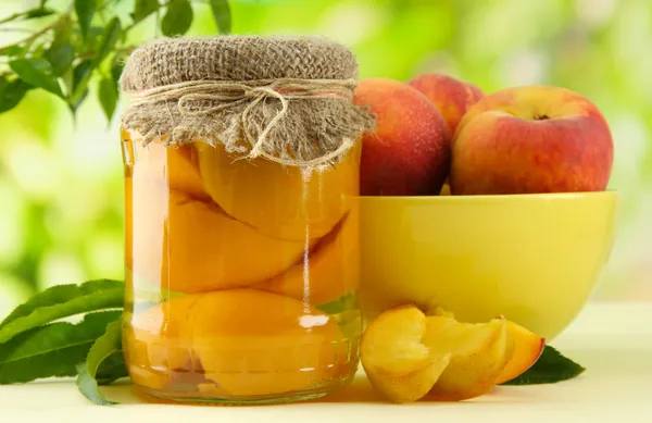 Jar of canned peaches and fresh peaches on wooden table, outside