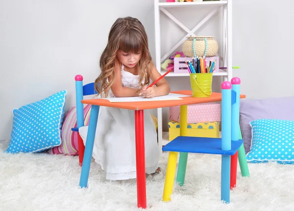 Little girl draws sitting at table in room on grey wall background