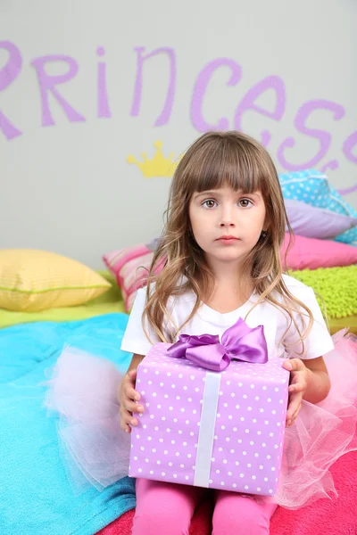 Little girl sitting on bed with gift in room on grey wall background