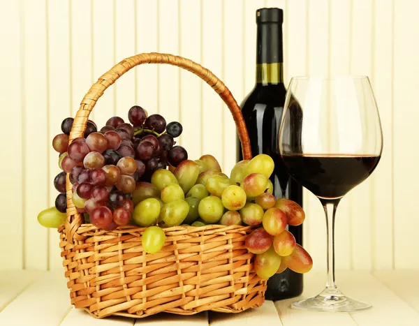 Ripe grapes in wicker basket, bottle and glass of wine, on light background