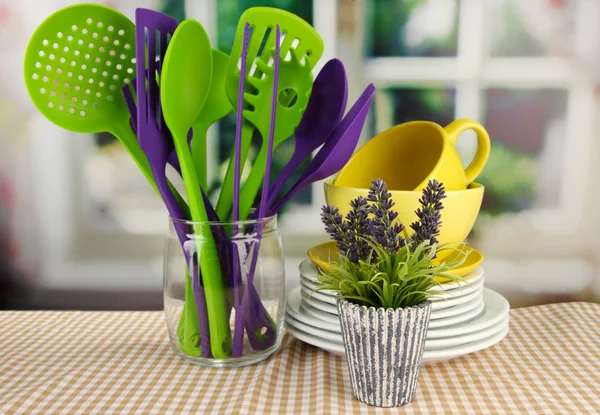 Plastic kitchen utensils in stand with clean dishes on tablecloth on bright background