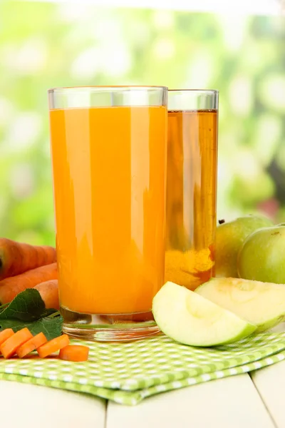 Glasses of juice, apples and carrots on white wooden table, on green background
