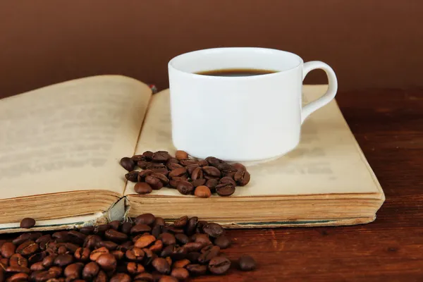 Cup of coffee with coffee beans and book on wooden table on brown background