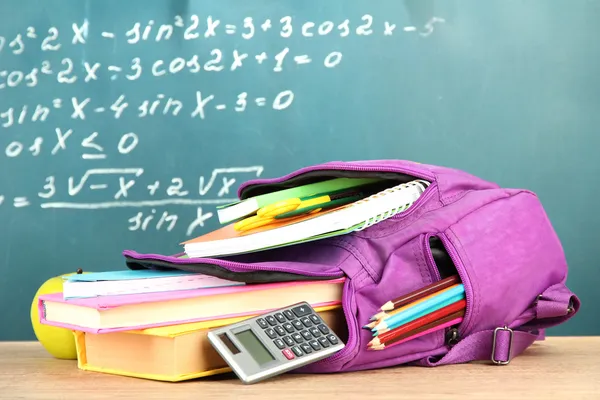 Purple backpack with school supplies on wooden table on green desk background