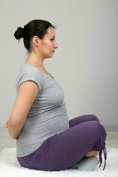 Young pregnant woman doing exercises on rug on wall background