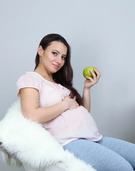 Young regnant woman sitting on armchair and holding apple on wall background