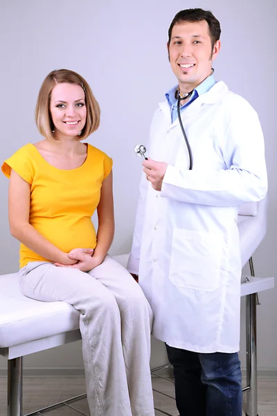 Young pregnant woman sitting on hospital bed with doctor on gray background