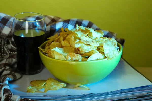 Chips in bowl, cola and TV remote on wooden table on colorful background