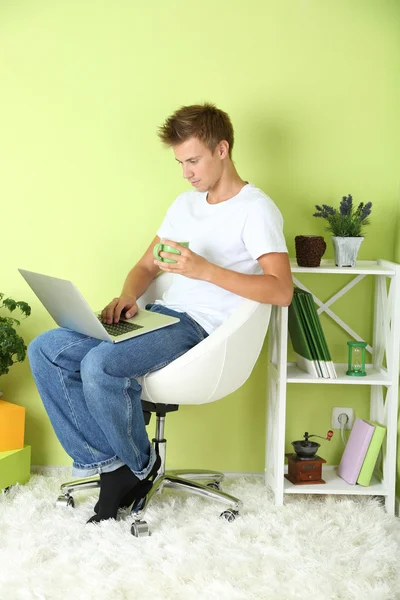Young man relaxing with laptop in armchair, on home interior background