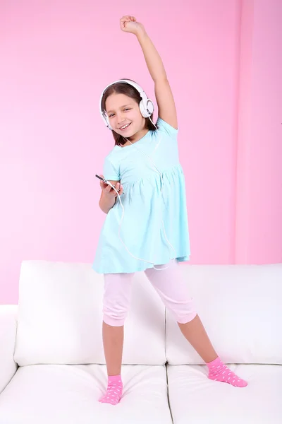Beautiful little girl listening to music and dancing on sofa and listening to music, on home interior background