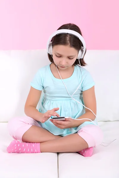 Beautiful little girl sitting on sofa and listening to music, on home interior background