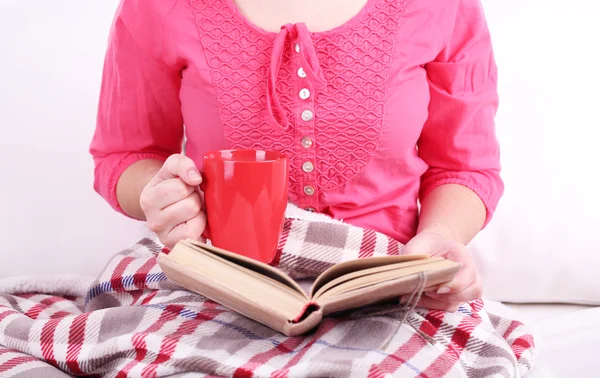 Woman sitting on sofa, reading book and drink coffee or tea, close-up