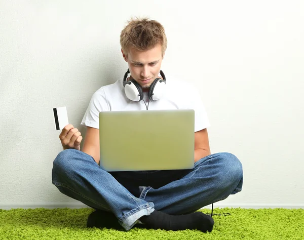 Young man sitting on carpet with laptop and credit card, on gray wall background