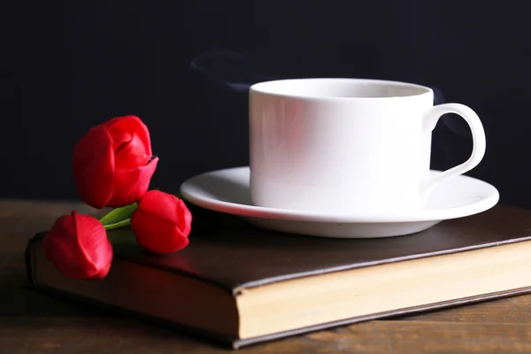 Cup of hot coffee on book with flowers on table on dark background