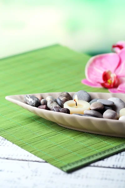 Spa stones and candle in decorative bowl, on wicker mat, on color wooden background