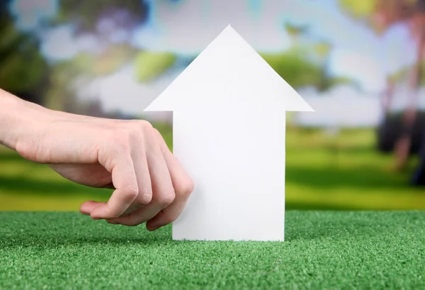 Little paper house in hand close-up, on green grass, on bright background