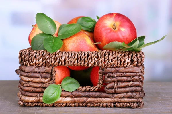 Ripe sweet apples with leaves in wicker crate on light background