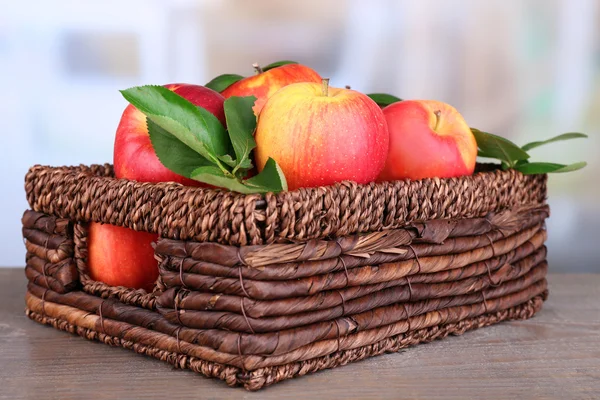 Ripe sweet apples with leaves in wicker crate on light background