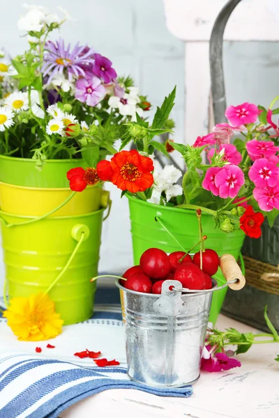 Bouquet of colorful flowers and fresh cherries in decorative buckets, on chair, on light wall background