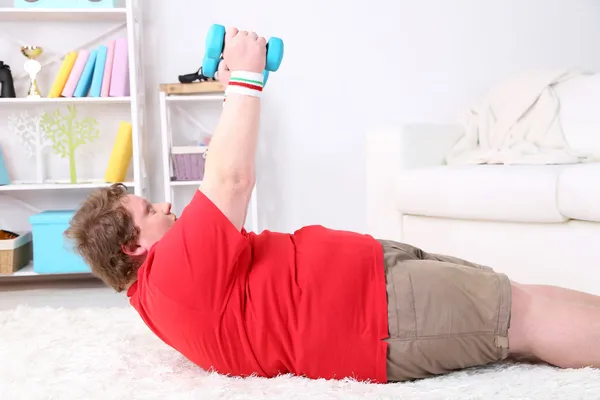 Large fitness man making exercises with dumbbells on floor, at home