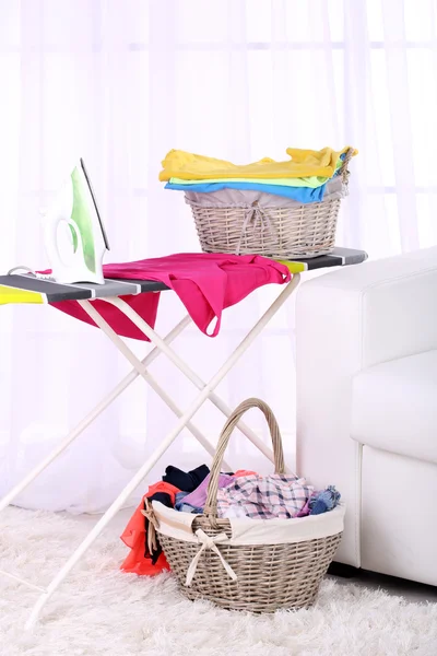 Baskets with laundry and ironing board on light home interior background
