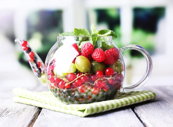 Forest berries and mint leaves in glass teapot, on wooden table, on bright background