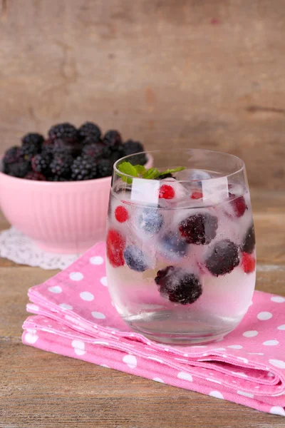 Cold cocktail with forest berries, frozen in ice cubes and fresh blackberry in bowl on color wooden background