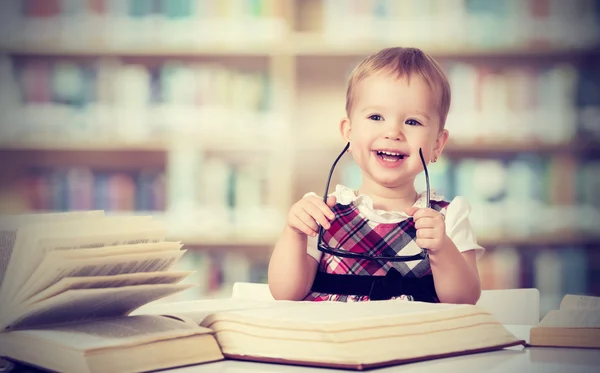 Funny baby girl in glasses reading a book in a library