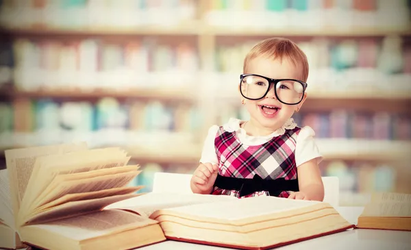 Funny baby girl in glasses reading a book in a library
