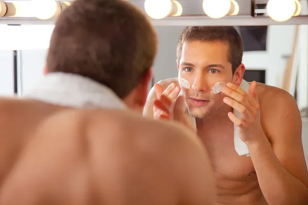 Reflection of young man in mirror applying shaving cream