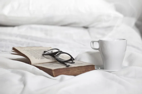 Closeup of a cup of coffe, old book and rimmed glasses on a white pillow.