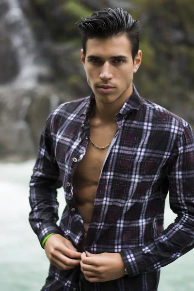 Handsome young man near mountain waterfall with open shirt
