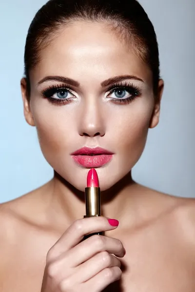 Glamor closeup portrait of beautiful sexy Caucasian brunette young woman model applying makeup lipstick on her lips with perfect clean skin