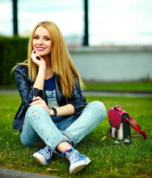 Portrait of cute funny blond modern sexy urban young stylish smiling woman girl model in bright modern cloth outdoors sitting in the park in jeans with pink bag