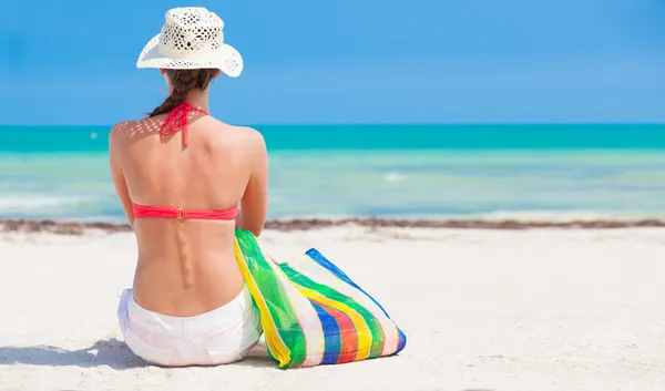 Woman in bikini and straw hat with beach bag sitting on beach. back view