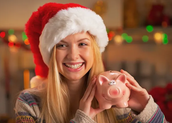 Teenage girl in santa hat showing piggy bank in christmas decorated kitchen
