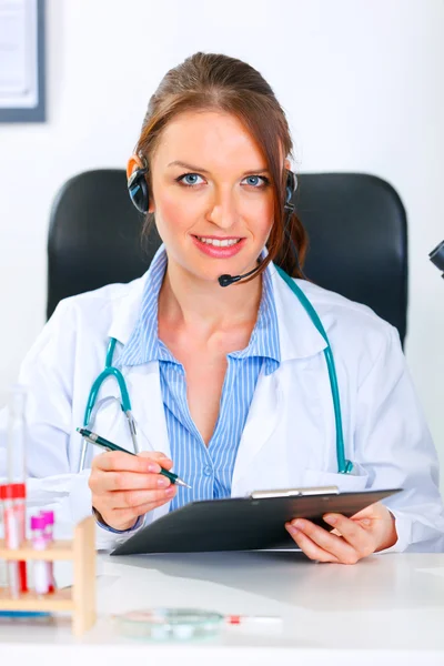 Smiling doctor woman with headset sitting at office table and holding clip