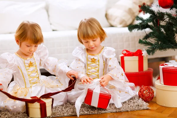 Two happy twins girl opening presents near Christmas tree